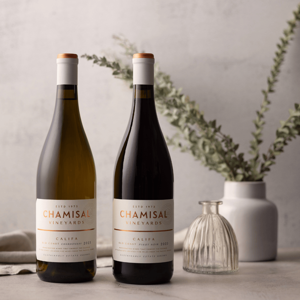 Two bottles from the Califa Chardonnay & Pinot Noir 2-Bottle Gift stand side by side on a light surface, with a white cloth and two vases of leafy branches behind them. The softly lit scene is elegant and minimalistic.