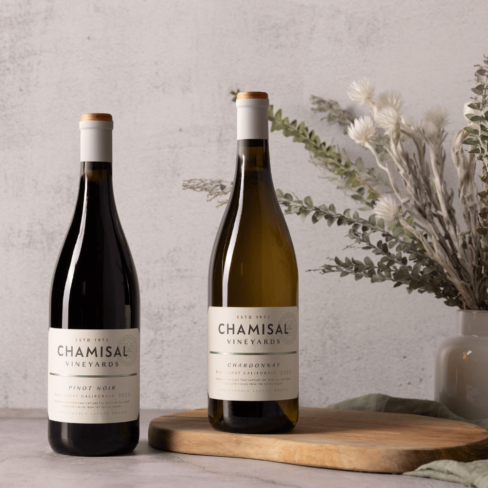 Two Chamisal Vineyards wine bottles, the Califa Chardonnay & Pinot Noir 2-Bottle Gift, are displayed on a table with a wooden board. A vase with dried flowers and foliage sits in the background.