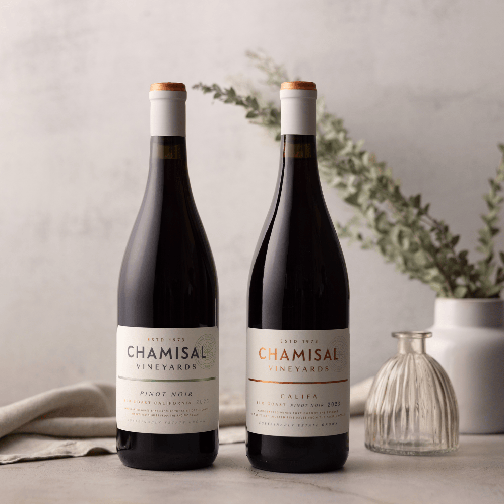 Two bottles from the Califa Chardonnay & Pinot Noir 2-Bottle Gift are displayed on a light surface with a soft, neutral background. A clear glass vase with greenery sits behind the bottles, adding a fresh touch to the elegant scene.
