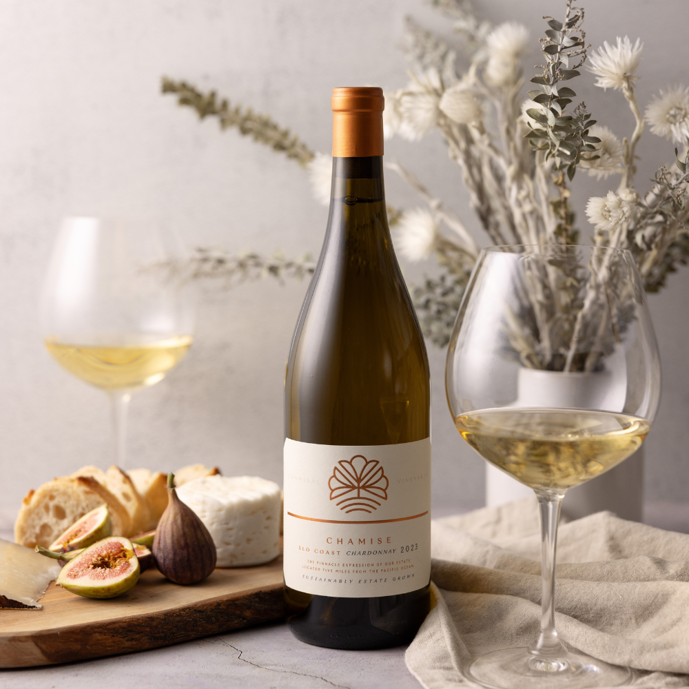 A bottle of white wine with two filled wine glasses sits on a table beside a cheese board with bread, cheese, figs, and pears. In the background, a vase holds dried white flowers and eucalyptus.