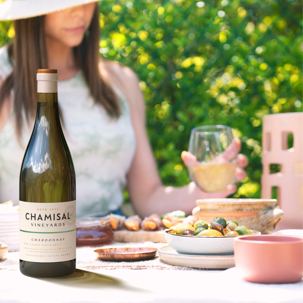 A bottle of Chamisal Vineyards Chardonnay sits on an outdoor table set with food and ceramics. In the background, a woman in a sunhat holds a glass of white wine, with greenery behind her.