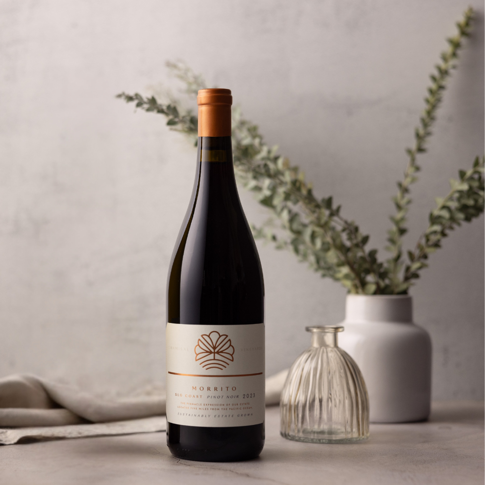 A bottle of Morrito Pinot Noir 2023 stands on a light surface with a white vase of greenery and a ribbed glass bottle blurred in the background, creating a minimalist and elegant setting.