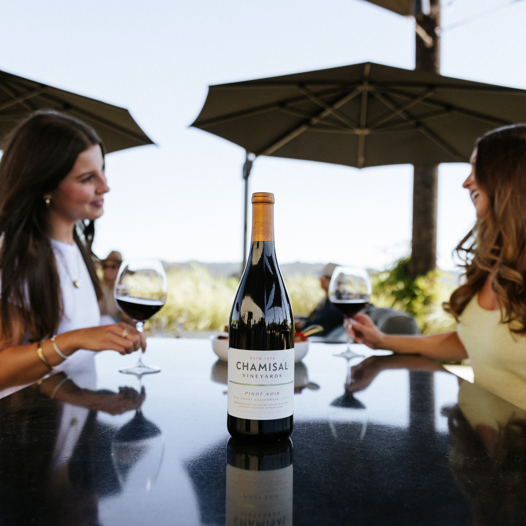 A bottle of Chamisal Vineyards Pinot Noir sits on a reflective table outdoors, with two women sitting across from each other, each holding a glass of red wine. Large umbrellas provide shade in the background.