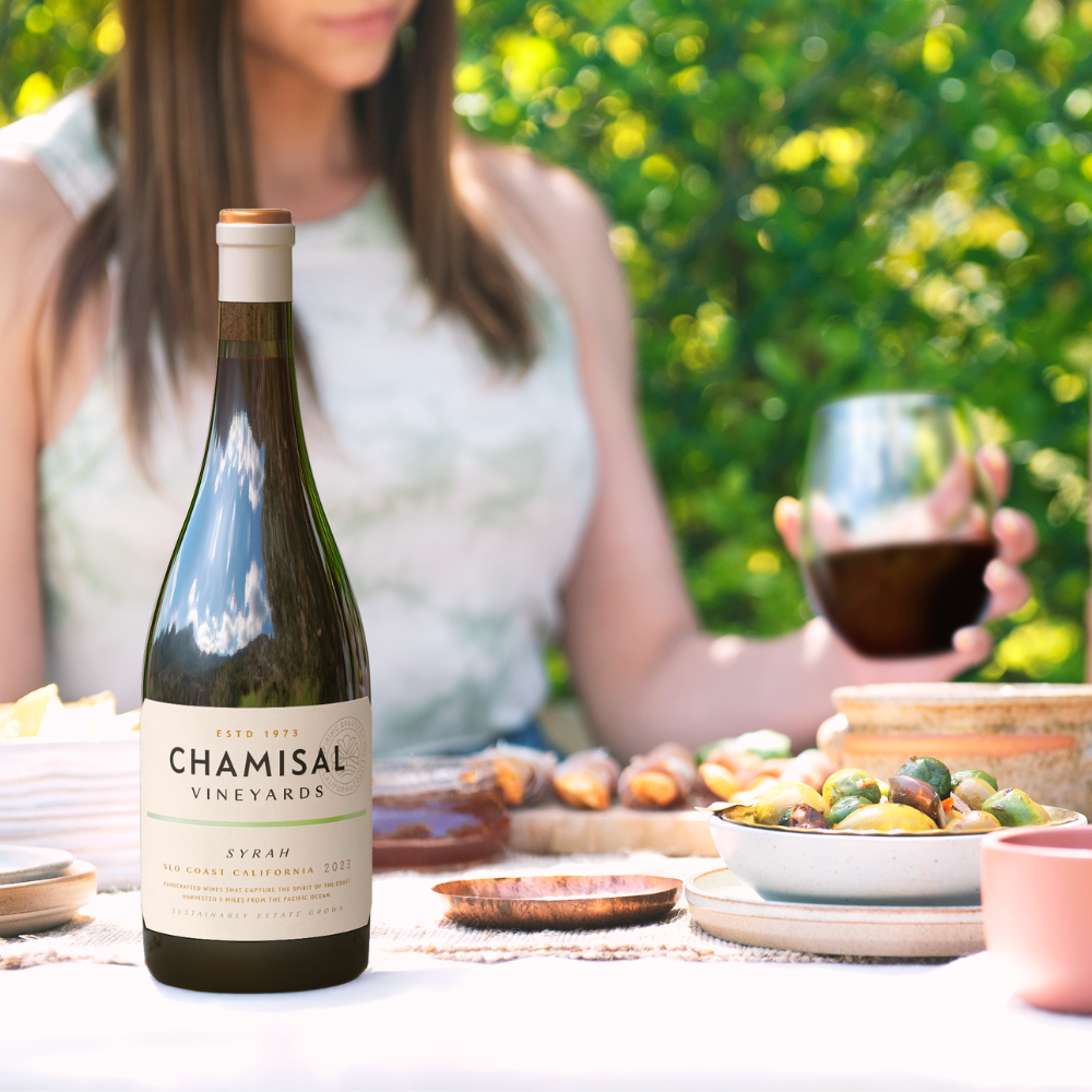 A bottle of Chamisal Vineyards Syrah stands on a table set with food, while a woman in a sleeveless top sits in the background holding a glass of red wine outdoors on a sunny day.
