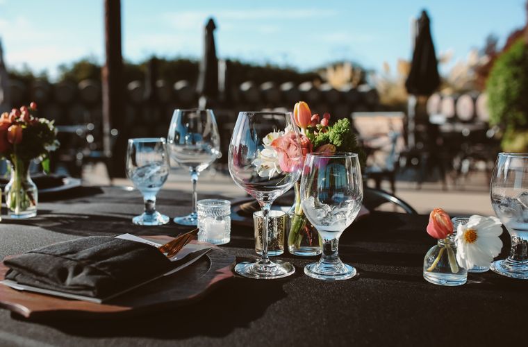 An elegant outdoor table setting with empty wine glasses, black napkins, small flower arrangements, and candles, set on a black tablecloth. Wine barrels and blurred outdoor seating are visible in the background.