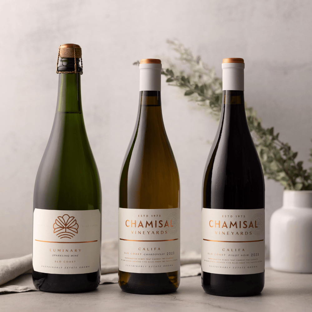 Three bottles from the Chamisal Holiday Entertaining 6-Bottle Collection—one sparkling, one white, and one red—stand side by side on a light surface with a minimal background and a ceramic vase with greenery.