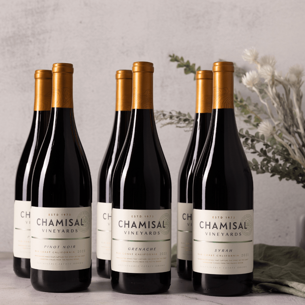 Seven bottles from the San Luis Obispo Coast Reds 6-Bottle Collection with gold foil tops are arranged on a light surface. Floral and leafy decorations in the background create an elegant display.