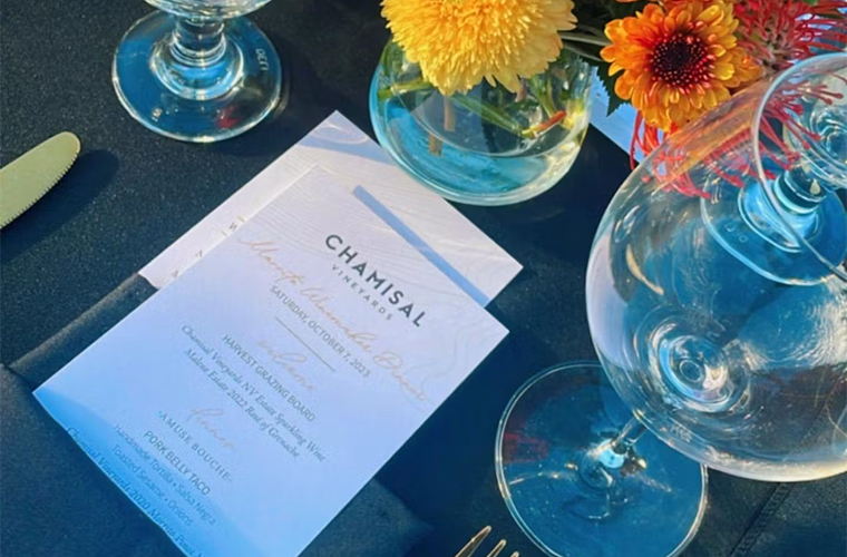 A close-up of a table set with a Morrito menu, wine glass, water glass, cutlery, and a small bouquet of yellow and orange flowers on a black tablecloth, with sunlight shining on the scene.
