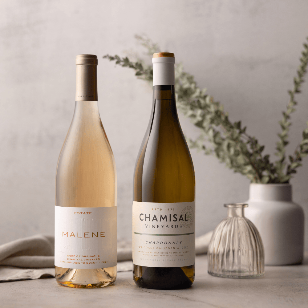 Two wine bottles stand side by side on a light surface: a Malene rosé on the left and a Chamisal Vineyards Chardonnay on the right—capturing the elegance of the Chamisal & Malene Duo Wine Gift. In the background are two vases, one with greenery.
