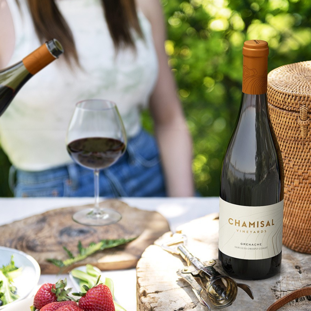 A bottle of Chamisal Vineyards wine sits on a picnic table with a wine glass, strawberries, and utensils. A person in a white top is in the background, and a woven basket is on the right, all set outdoors in greenery.