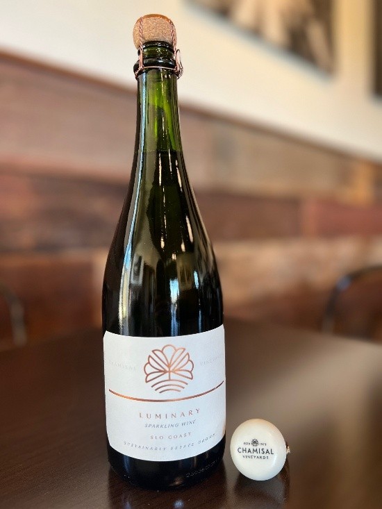 A bottle of Luminary Sparkling Wine from Chamisal Vineyards sits on a dark table, its cork and wire cage nearby—an inviting scene reminiscent of those featured in wine club newsletters. Wood paneling and blurred wall images provide a cozy backdrop.