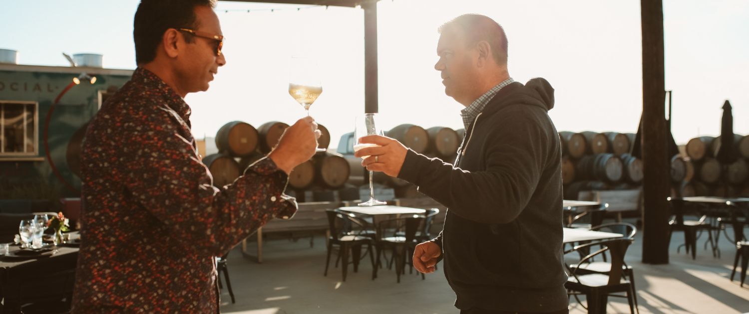 Two men stand outdoors at a winery, clinking wine glasses. Wine barrels and tables are visible in the background, with sunlight streaming in, creating a warm atmosphere—perfect for a feature in wine club newsletters.