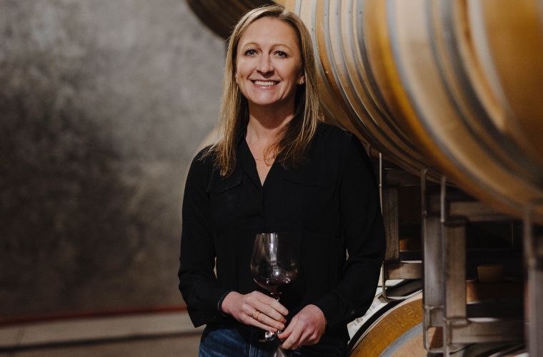 A woman with long blonde hair, wearing a black blouse and jeans, stands indoors next to large wine barrels, smiling and holding a glass of red wine—perhaps celebrating her latest wine club selection.