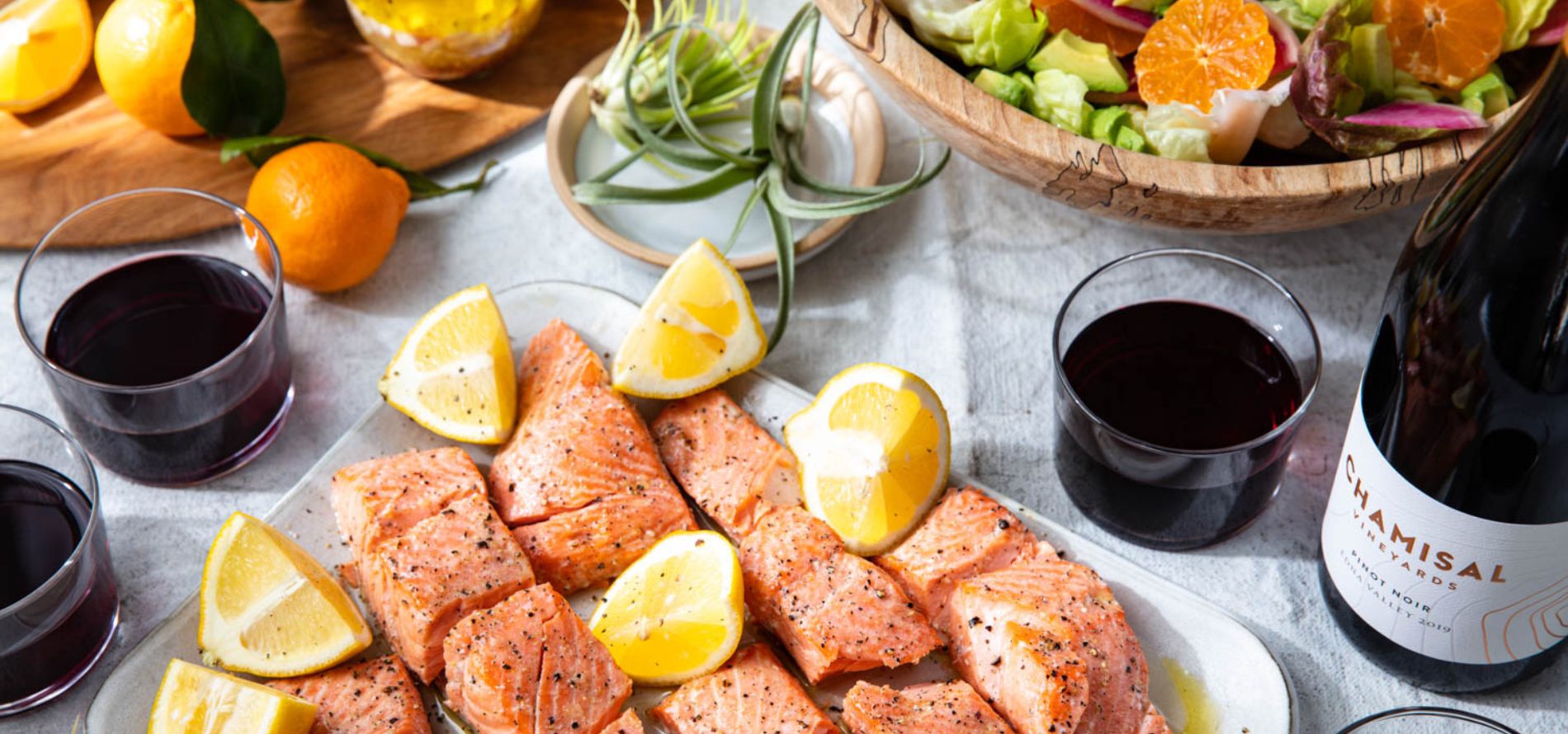A table set with grilled salmon pieces garnished with lemon wedges, glasses of red wine, a bottle of wine, a bowl of salad with oranges, tangerines, olive oil, and fresh green plants as decoration.