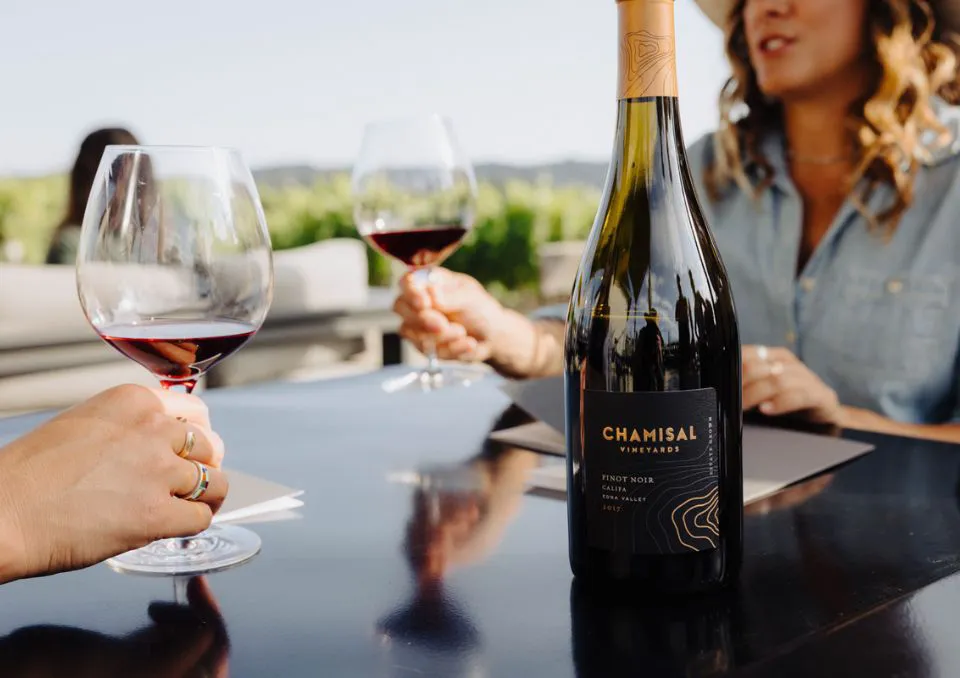 Two people sit at an outdoor table, each holding a glass of red wine. A bottle of Chamisal Vineyards Pinot Noir sits in the foreground, with vineyards visible in the background.