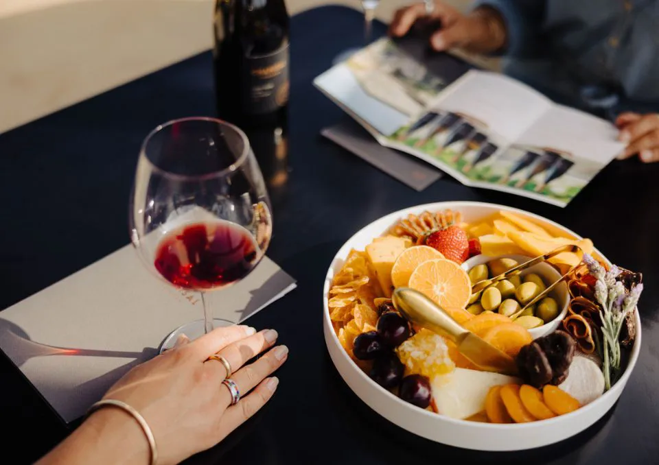 A person’s hand holding a glass of red wine sits near a platter of assorted cheeses, fruits, nuts, and olives on a dark table, with another person reading a book in the background.