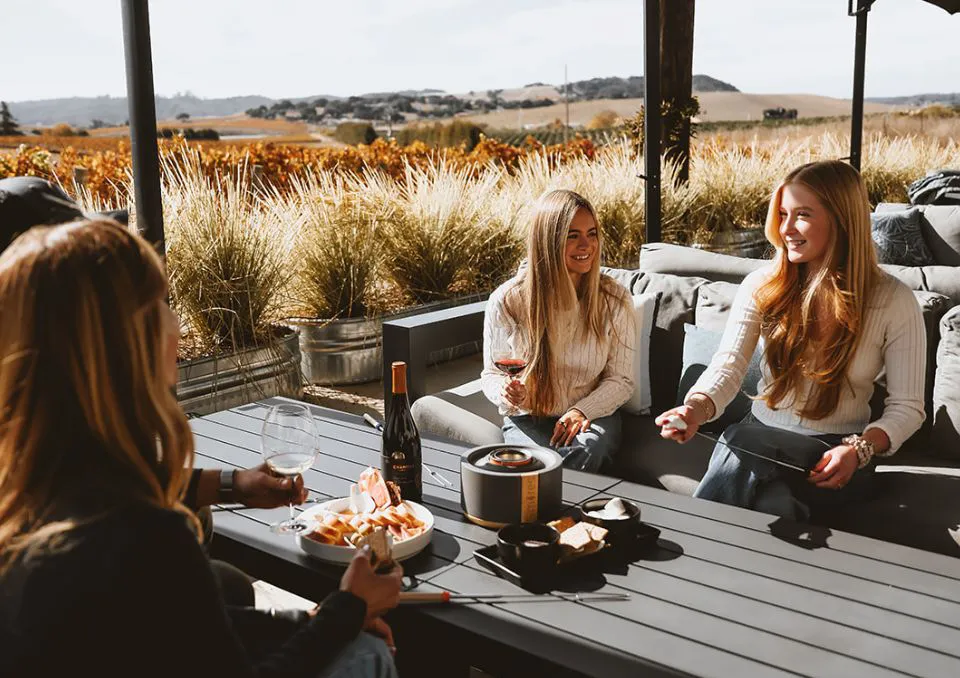 Three women sit at an outdoor table with wine and snacks, smiling and talking, with a scenic vineyard and hills in the background on a sunny day.
