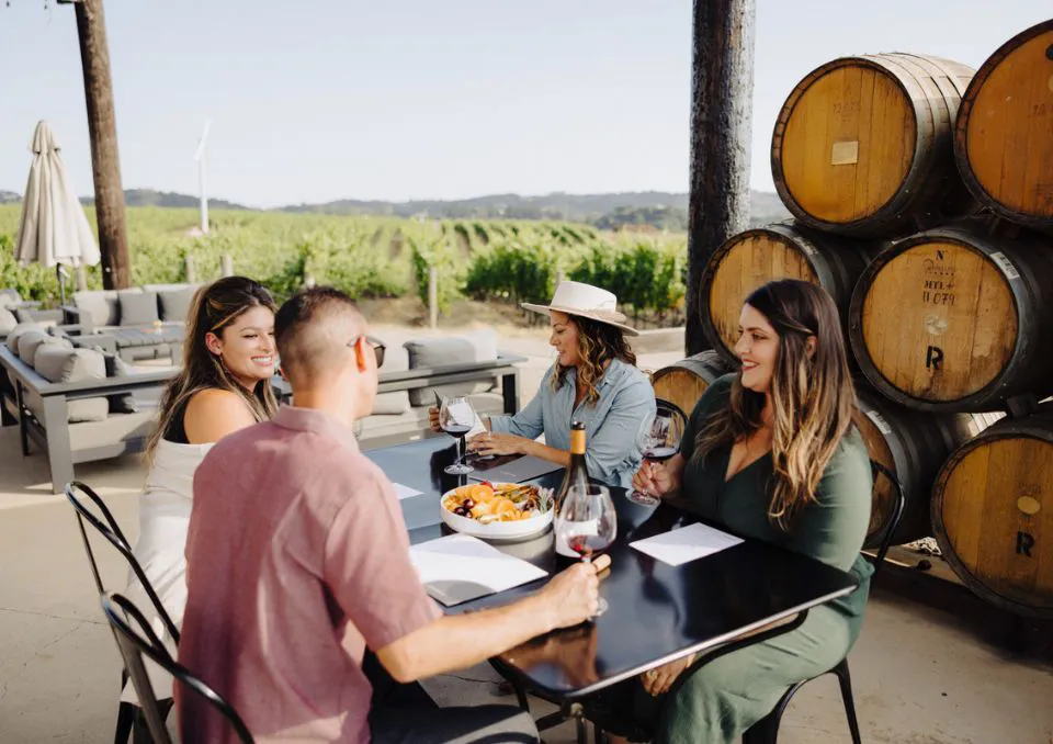 Four people sit at an outdoor table near wine barrels, enjoying wine and snacks. They are smiling and talking, with a vineyard and patio seating visible in the background.