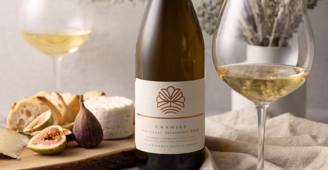 A bottle of Chamise Chardonnay 2020 is placed on a table with two glasses of white wine, a wooden board with bread, figs, and cheese, and a vase with dried flowers in the background.