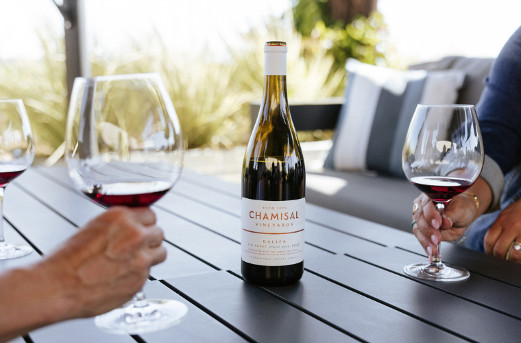 A bottle of Chamisal Vineyards wine sits on an outdoor table, surrounded by three people holding glasses of red wine. The setting appears relaxed and sunny, with blurred greenery in the background.
