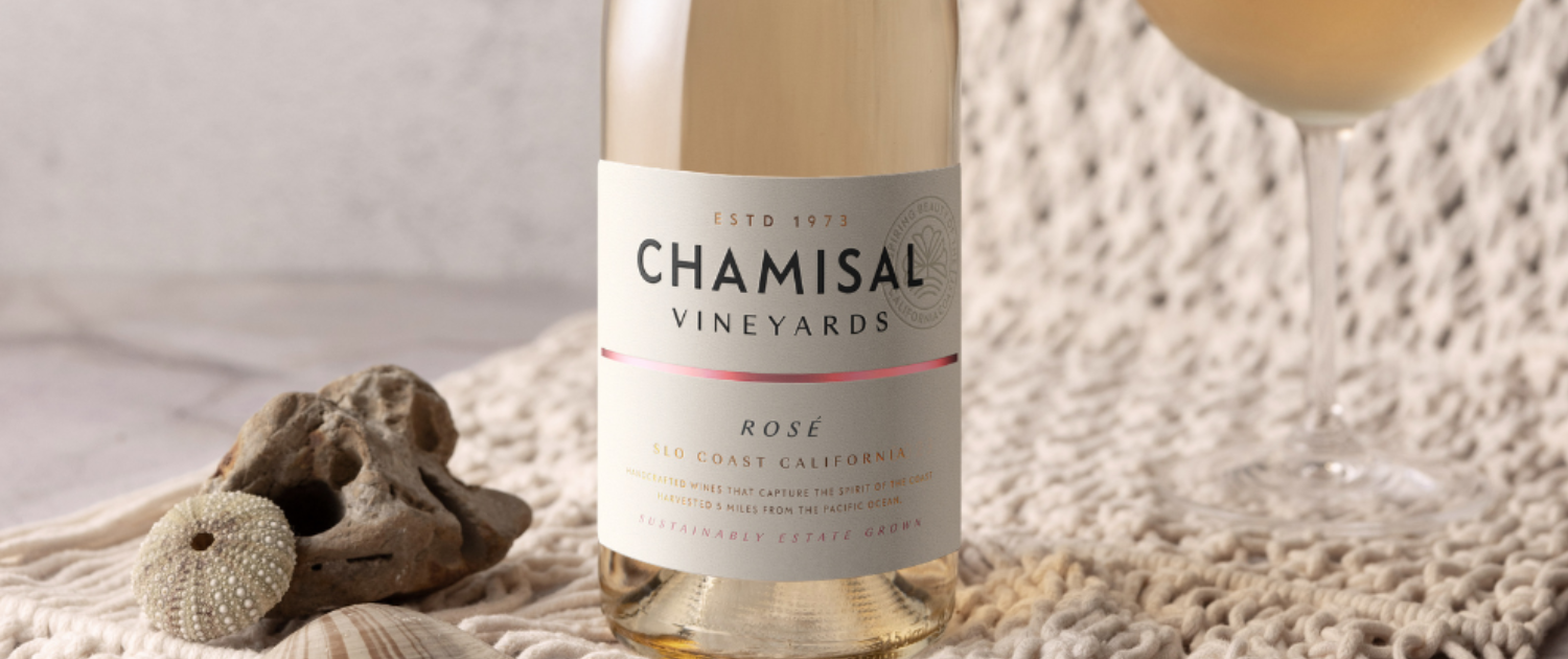 A bottle of Chamisal Vineyards Rosé wine sits on a textured cream-colored cloth next to seashells, evoking the elegance often featured in wine club newsletters, with a filled wine glass partially visible in the background.