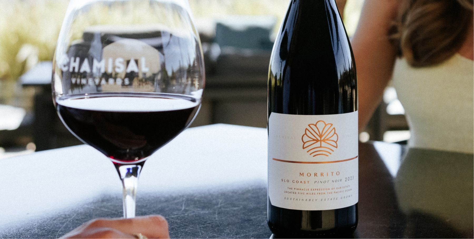 A close-up of a glass of red wine and a bottle labeled “Morrito SLO Coast Pinot Noir 2021” on a table, with blurred people in the background and outdoor lighting, showcases one of the best Edna wineries in a vibrant setting.