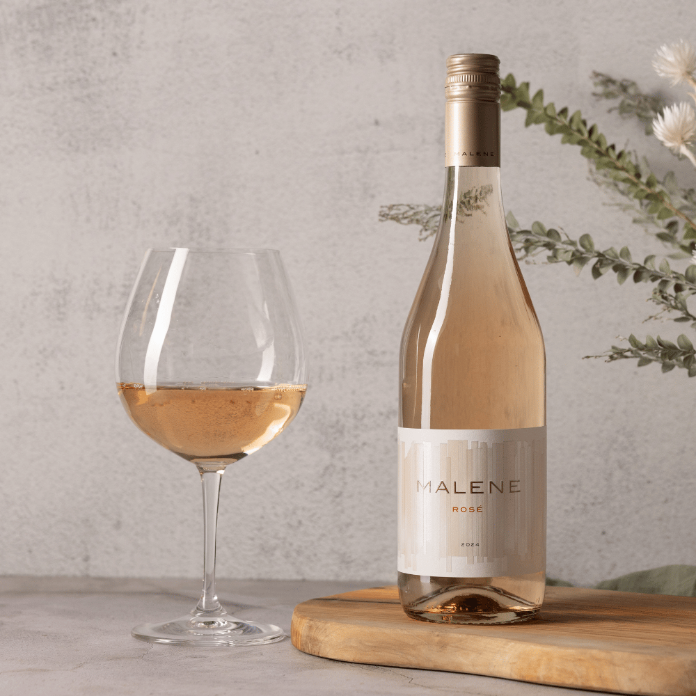 A bottle of Malene Rosé from the Entertaining 6-Bottle Collection stands next to a filled wine glass on a wooden board, with green foliage and white flowers in the background against a light gray textured wall.
