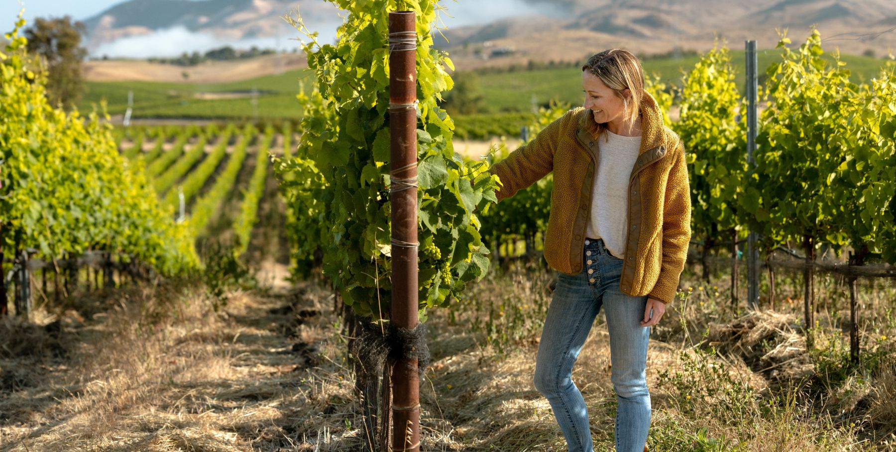 A woman in a yellow jacket and jeans smiles while touching a grapevine in a vineyard, with rows of green vines and hills in the background under a partly cloudy sky.
