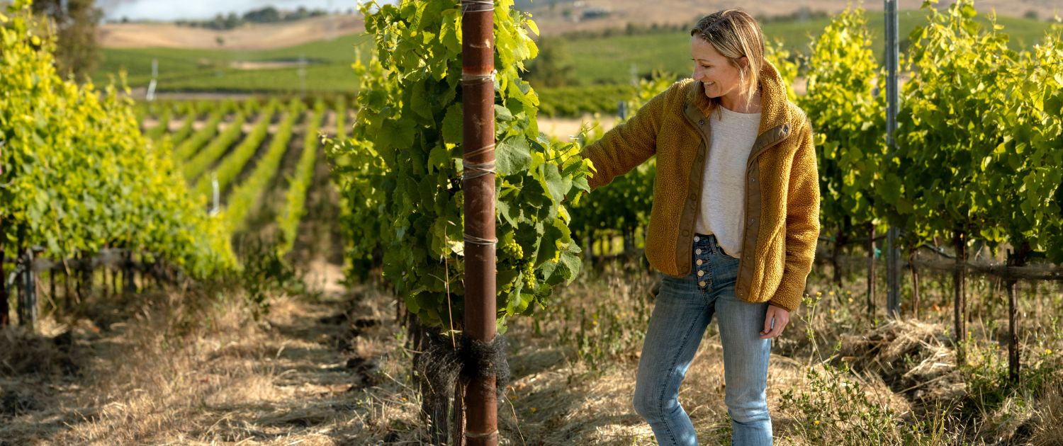 A woman in a yellow sweater and jeans smiles while touching grapevines in a vineyard on a sunny day, surrounded by rows of green plants and scenic hills in the background—a perfect scene often featured in wine club newsletters.