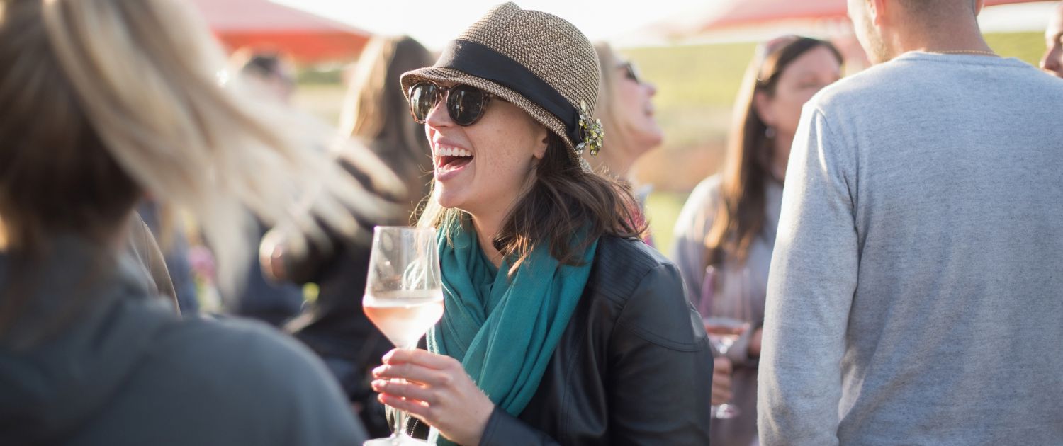 A woman wearing a hat, sunglasses, and a teal scarf laughs while holding a glass of wine outdoors, surrounded by people socializing in bright, natural light—capturing the lively spirit often featured in wine club newsletters.
