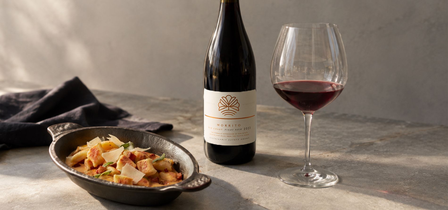 A bottle of red wine, a glass of red wine, and a dish of pasta with shaved cheese are set on a stone surface in soft natural light.