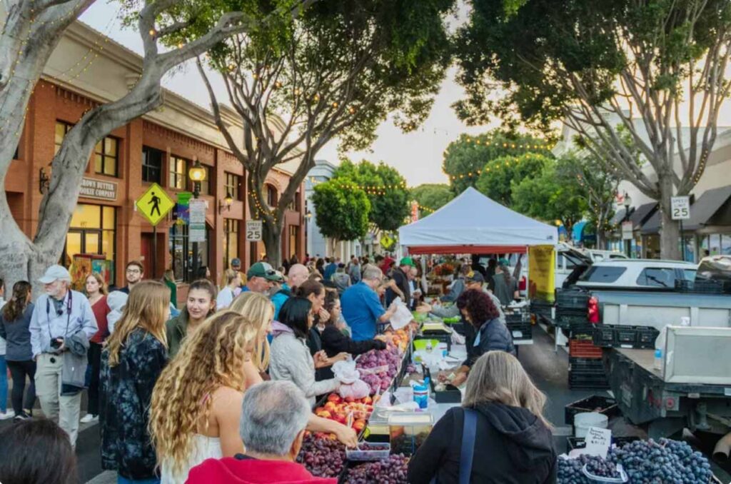 People shop for fresh produce at an outdoor farmers market lined with tables of fruits and vegetables, surrounded by trees and brick buildings, under a canopy of string lights.