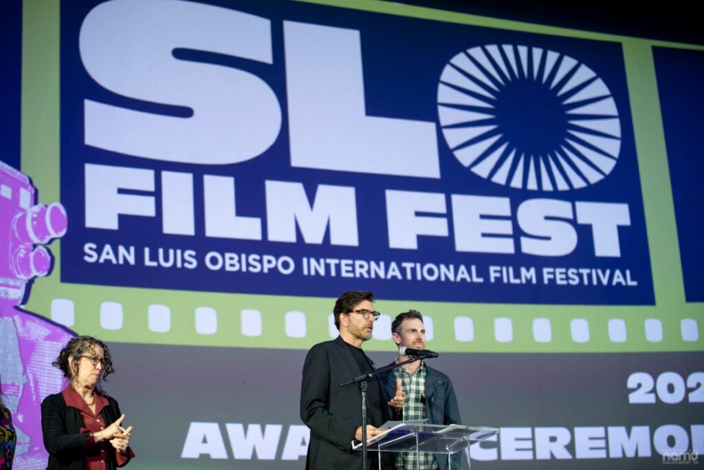 Three people stand on stage at a podium during the SLO Film Fest awards ceremony. A large event banner is displayed behind them with the festival name and year.