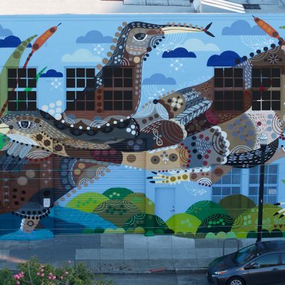 A colorful mural on a building features a large stylized bird made of geometric shapes and patterns, surrounded by clouds and green hills, with windows incorporated into the design. A car is parked on the street below.