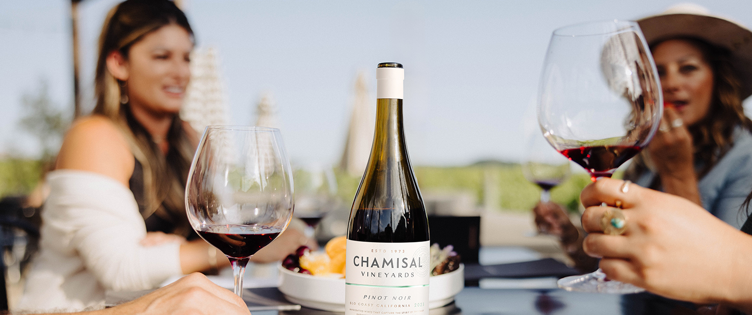 A bottle of Chamisal Vineyards Pinot Noir centered on a table between two people holding glasses of red wine at a vineyard, the perfect scene for wine club newsletters.
