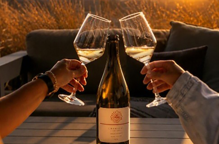 Two people in a wine club toasting with glasses of white wine in front of a wine bottle at sunset.